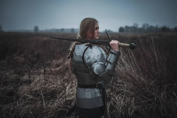 Menina na imagem de Jeanne d 'Arc em armadura e com espada em suas mãos fica no prado. Visão traseira . — Fotografia de Stock