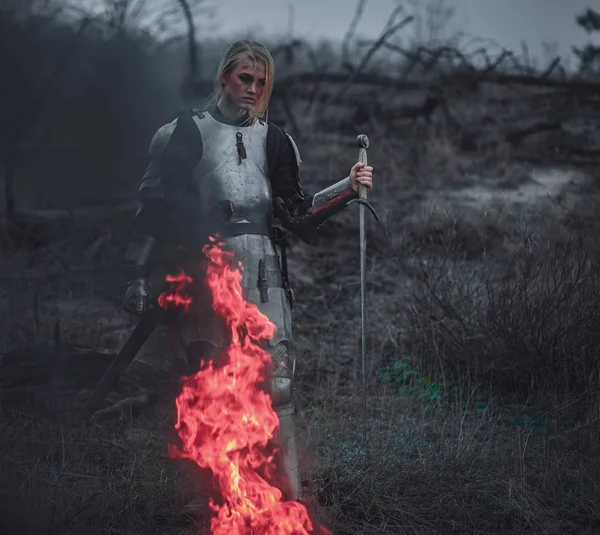 Menina na imagem de Jeanne d 'Arc em armadura e com espada em suas mãos fica contra o fundo de fogo e fumaça . — Fotografia de Stock