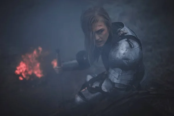 Chica en la imagen de Jeanne d 'Arc en armadura y con la espada en las manos se arrodilla contra el fondo de fuego y humo . — Foto de Stock
