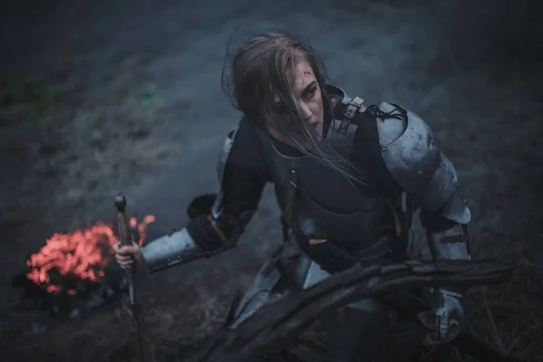 Fille à l'image de Jeanne d'Arc en armure et avec l'épée dans les mains s'agenouille sur fond de feu et de fumée . — Photo