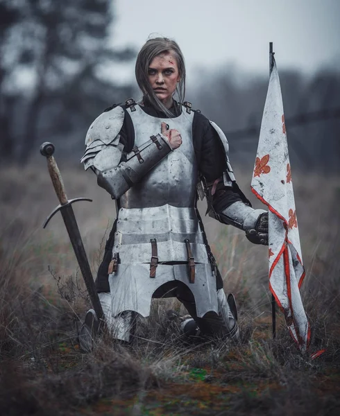 Chica en la imagen de Jeanne d 'Arc en armadura se arrodilla con la bandera en sus manos y espada en el prado . — Foto de Stock