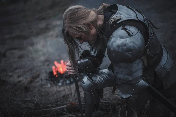 Ragazza a immagine di Giovanna d'Arco si inginocchia in armatura e con la spada tra le mani sullo sfondo di fuoco e fumo . — Foto Stock