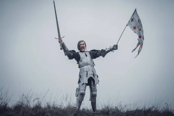 Chica en la imagen de Jeanne d 'Arc está de pie en armadura y emite grito de batalla con la espada levantada y la bandera en sus manos . — Foto de Stock