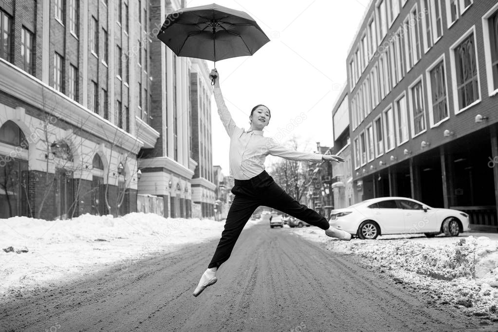 Ballerina with umbrella in her hands is high jumping at city street. Black and white image.