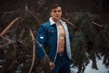 Meme bared ile açılarak ceketli genç kaslı adam kış ormanda çam ağacı yanında duruyor.