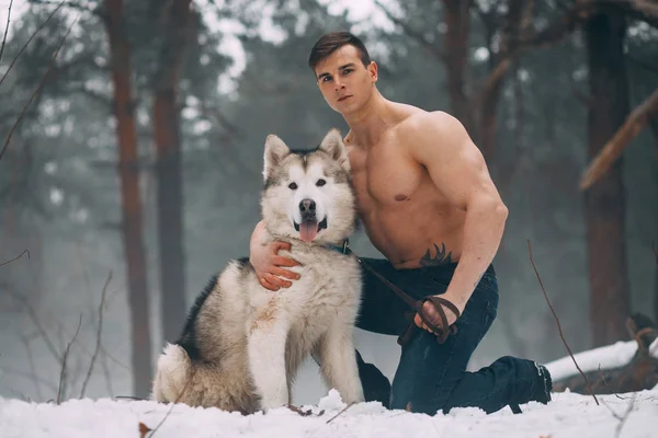 Young bodybuilder with bare torso embraces dog Malamute at walk in winter forest. — Stock Photo, Image