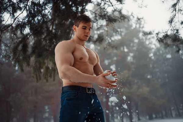 Young bodybuilder with bare torso wipes his body by snow in forest in winter.