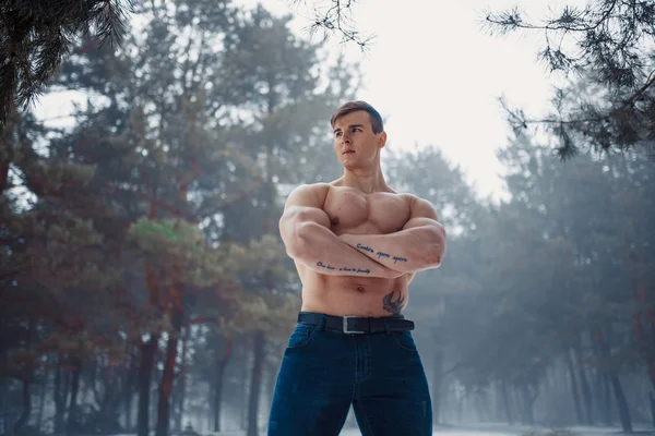 Young bodybuilder with bare torso stands with arms crossed in winter misty forest. — Stock Photo, Image