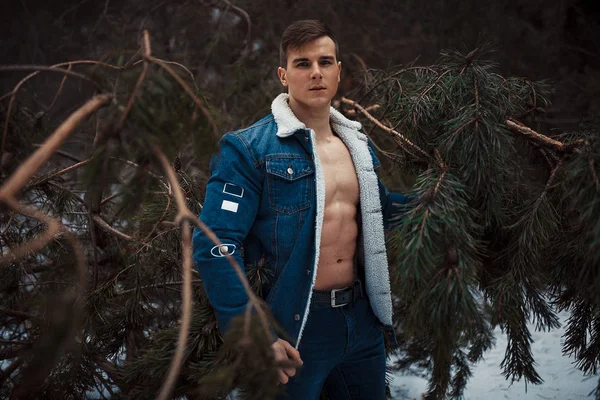 Young muscular man in unbuttoned jacket with bared breast stands next to pine tree in winter forest.