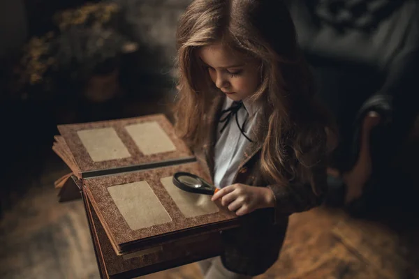 Gyermek lány a Sherlock Holmes képe áll a szobában és úgy néz ki photoalbum nagyítóval a háttérben a régi belső. Vértes. — Stock Fotó