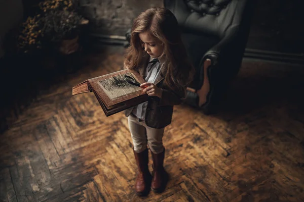 Menina na imagem de Sherlock Holmes fica no quarto e olha photoalbum com lupa no fundo do velho interior . — Fotografia de Stock