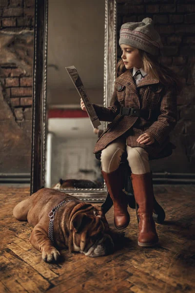 Niña en la imagen de Sherlock Holmes se sienta al lado del bulldog inglés y lee el periódico en el fondo del antiguo interior . — Foto de Stock