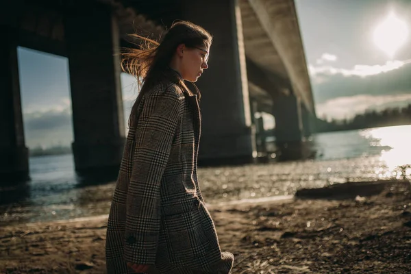 Jovem mulher caminha na margem do rio ao pôr do sol ao lado da ponte . — Fotografia de Stock