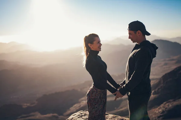 Couple se tient debout, tient la main et se regarde dans le contexte du lever du soleil dans les montagnes . — Photo