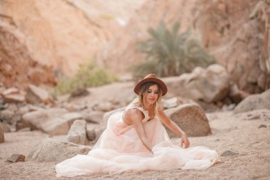 Gelin uzun elbise ve şapka kum kanyonda oturuyor.