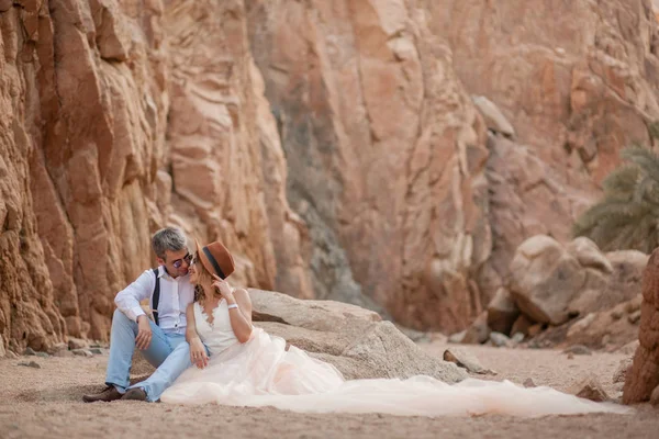 Novia y novio se sientan y sonríen en el cañón sobre el fondo de las rocas . —  Fotos de Stock