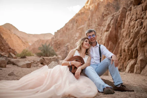 La novia y el novio se sientan y sonríen en el cañón sobre la arena sobre el fondo de las rocas . — Foto de Stock