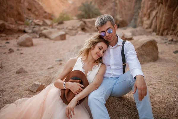 La novia y el novio se sientan y sonríen en el cañón sobre la arena sobre el fondo de las rocas. Primer plano. . —  Fotos de Stock
