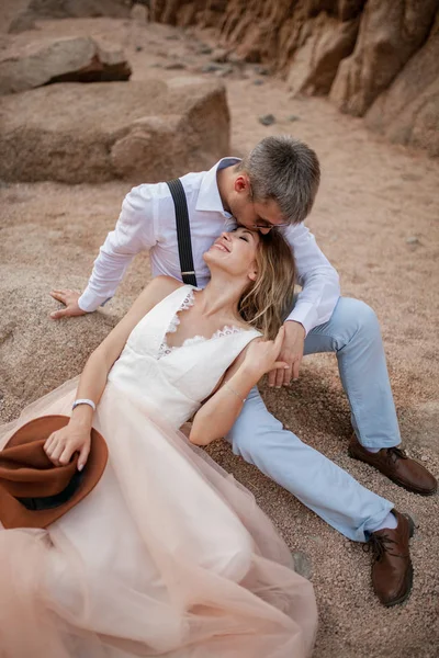 La novia y el novio se sientan y sonríen en la arena en el cañón sobre el fondo de las rocas. Primer plano. . — Foto de Stock