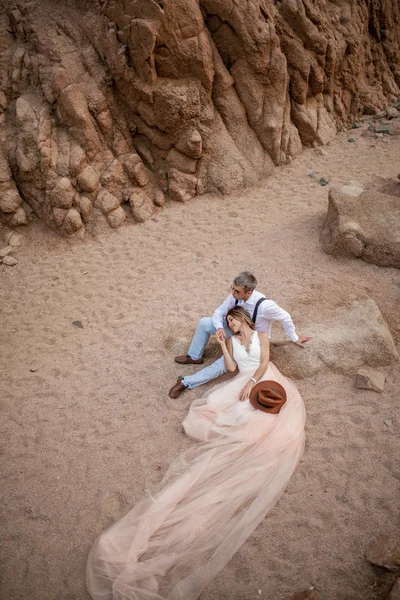 A noiva no vestido longo e o noivo sentam-se e mantêm mãos no cânion na areia. Vista superior . — Fotografia de Stock