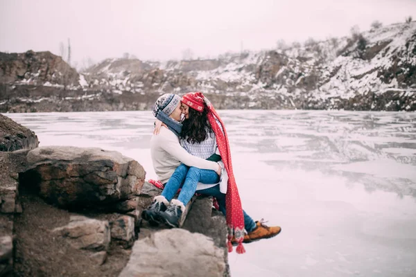 Paar zit bij rock, omarmen en glimlachend tegen een achtergrond van besneeuwde heuvels en bevroren meer. — Stockfoto