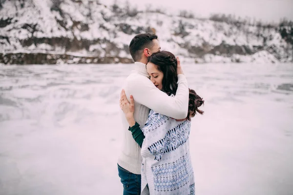 Pareja joven se está abrazando contra el fondo de colinas cubiertas de nieve y lago congelado . —  Fotos de Stock