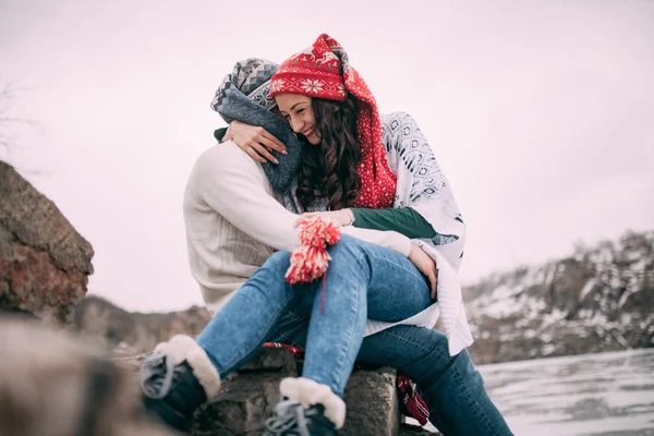 Das Paar sitzt am Fels, umarmt und lächelt vor dem Hintergrund schneebedeckter Hügel und des zugefrorenen Sees. Nahaufnahme. — Stockfoto