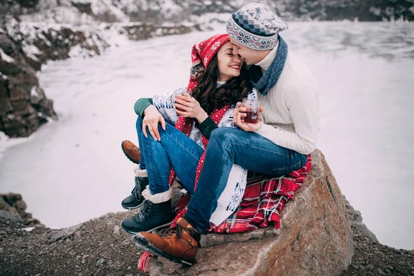 Young couple with cups of hot tea in their hands is sitting at rock and smiling during winter walk. — Stock Photo, Image