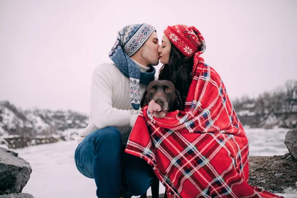 Junges Paar mit Hund im roten Karo sitzt und küsst sich beim Winterspaziergang. — Stockfoto