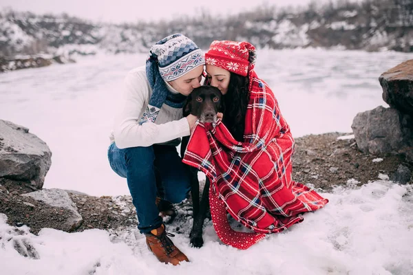 Pareja joven envuelta en cuadros rojos están sentados y besándose de perro durante el paseo de invierno . — Foto de Stock