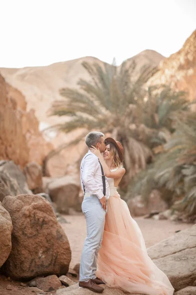 La novia y el novio se paran y se abrazan en el cañón contra el fondo de rocas y palmeras . — Foto de Stock