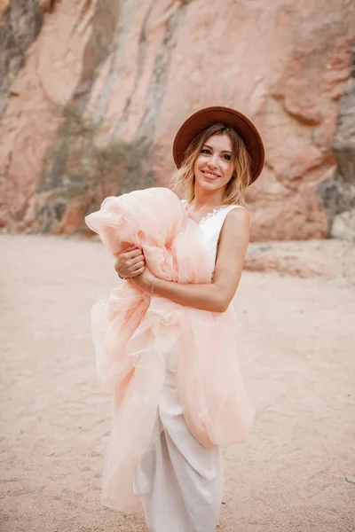 微笑的新娘在帽子站立和举行下摆她的长的礼服在峡谷. — 图库照片