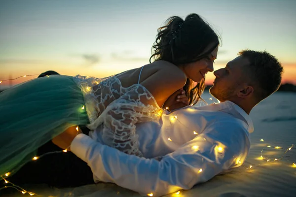 Mariés et mariés se trouvent dans le désert à côté de guirlande d'ampoules au coucher du soleil. Gros plan . — Photo