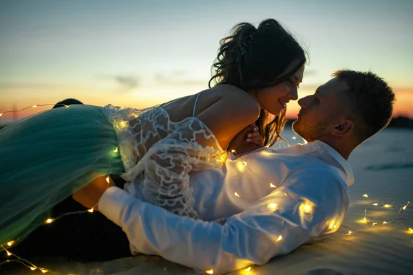 La novia y el novio se encuentran en el desierto junto a la guirnalda de bombillas al atardecer. Primer plano. . —  Fotos de Stock