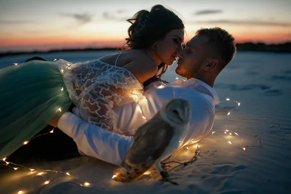 Noiva e noivo jazem no deserto e beijam ao lado da grinalda de lâmpadas e coruja ao pôr do sol. Fechar. . — Fotografia de Stock