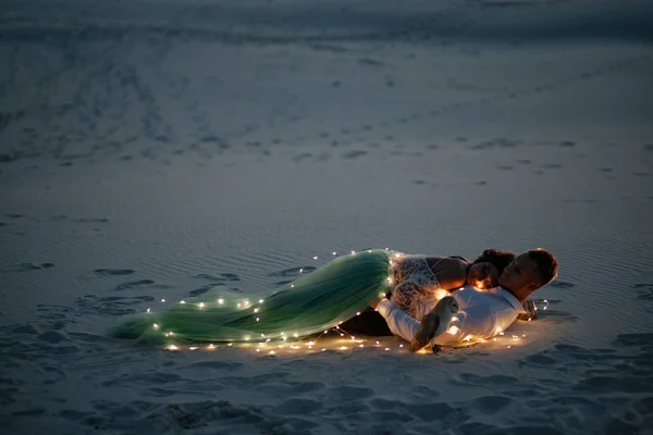 Noiva e noivo jazem no deserto ao lado de lâmpadas e coruja no fundo de pegadas na areia . — Fotografia de Stock