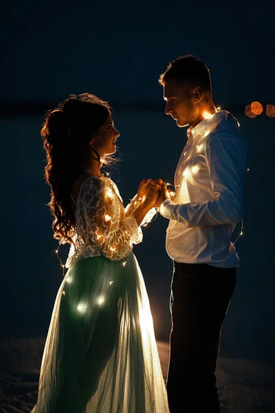 La novia y el novio se ponen de pie y se toman de la mano junto a la guirnalda de bombillas por la noche. Primer plano. . —  Fotos de Stock