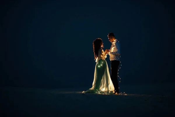 花嫁および新郎スタンド、手を握り合うと互いを見て砂漠の夜に電球のガーランド ・次へ. — ストック写真