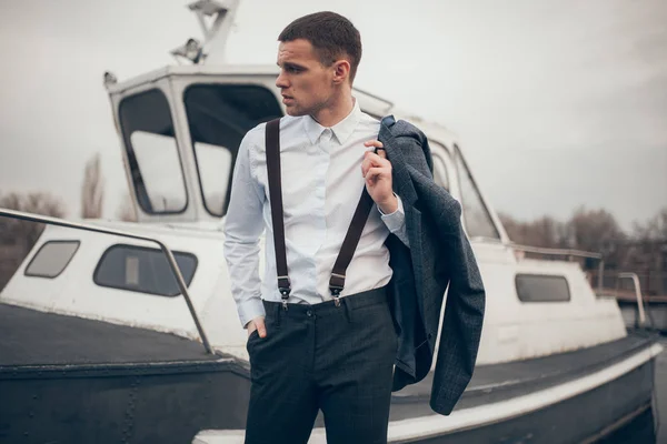 Молодий чоловік з курткою в руці стоїть на пірсі поруч з човном . — стокове фото