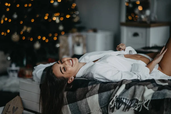 Mujer joven yace en la cama y sonríe sobre el fondo del árbol de Navidad — Foto de Stock