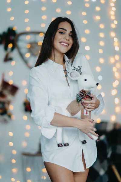 Щаслива жінка стоїть, тримає іграшкового оленя і посміхається на тлі різдвяних прикрас.. — стокове фото