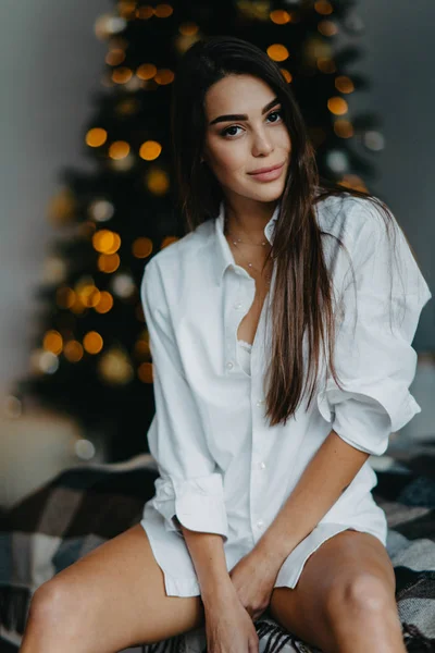 Mujer joven se sienta en la cama sobre el fondo del árbol de Navidad . —  Fotos de Stock