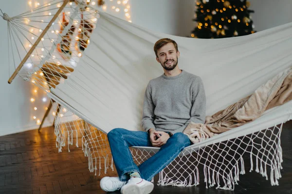 El hombre feliz se sienta en la hamaca contra el fondo del árbol de Navidad — Foto de Stock