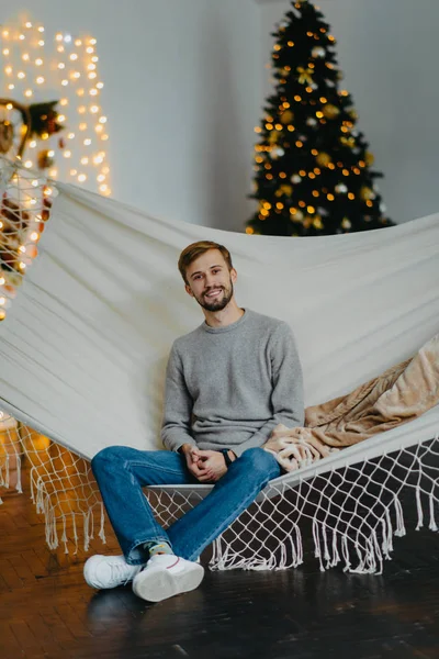 El hombre feliz se sienta en la hamaca contra el fondo del árbol de Navidad . — Foto de Stock