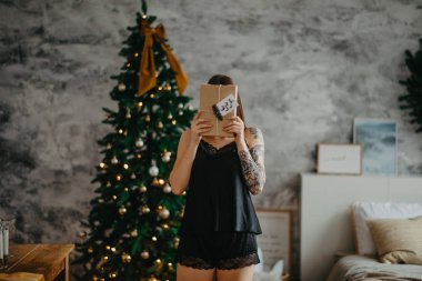 Siyah iç çamaşırlı genç bir kadın Noel ağacının yanında bir hediye kutusuyla duruyor..