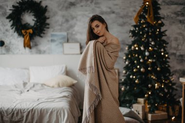 Ekoseye sarılı genç bir kadın Noel ağacının yanında duruyor..