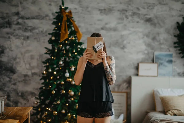 Νεαρή γυναίκα με μαύρα εσώρουχα στέκεται με ένα κουτί δώρου δίπλα στο χριστουγεννιάτικο δέντρο. — Φωτογραφία Αρχείου