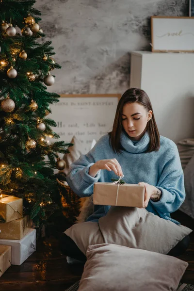 젊은 여자가 크리스마스 트리 옆에 앉아서 선물 상자를 엽니다. — 스톡 사진