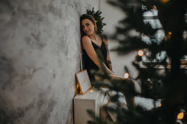 Fiatal nő fekete fehérnemű ül az ágyon mellett karácsonyfa — Stock Fotó