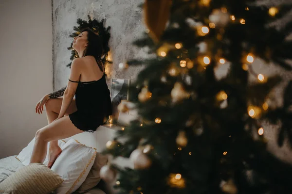 Mujer joven en lencería negra se sienta en la cama junto al árbol de Navidad — Foto de Stock